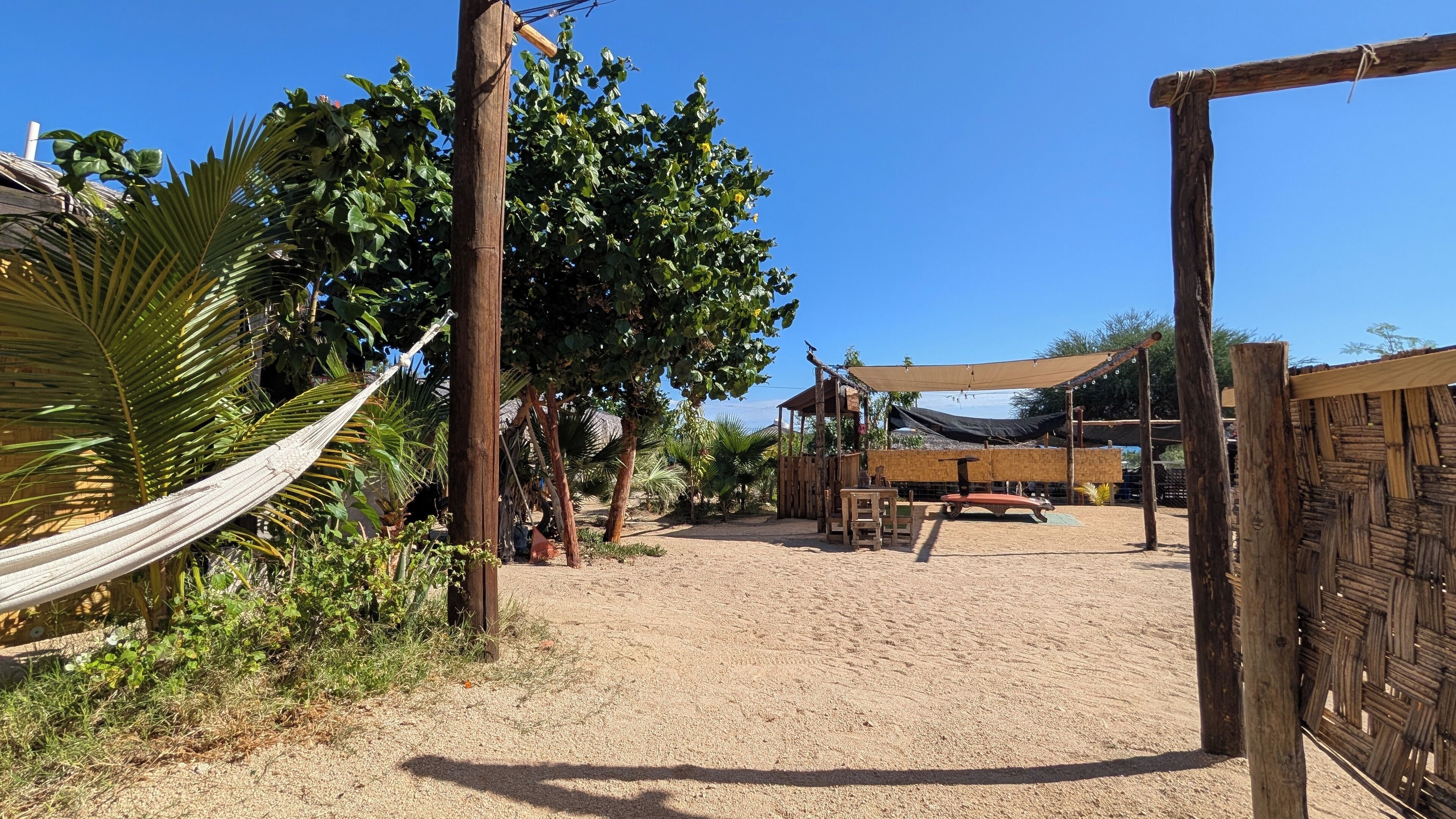Un espacio al aire libre con arena, una hamaca, estructuras de madera y un área de descanso a la sombra.