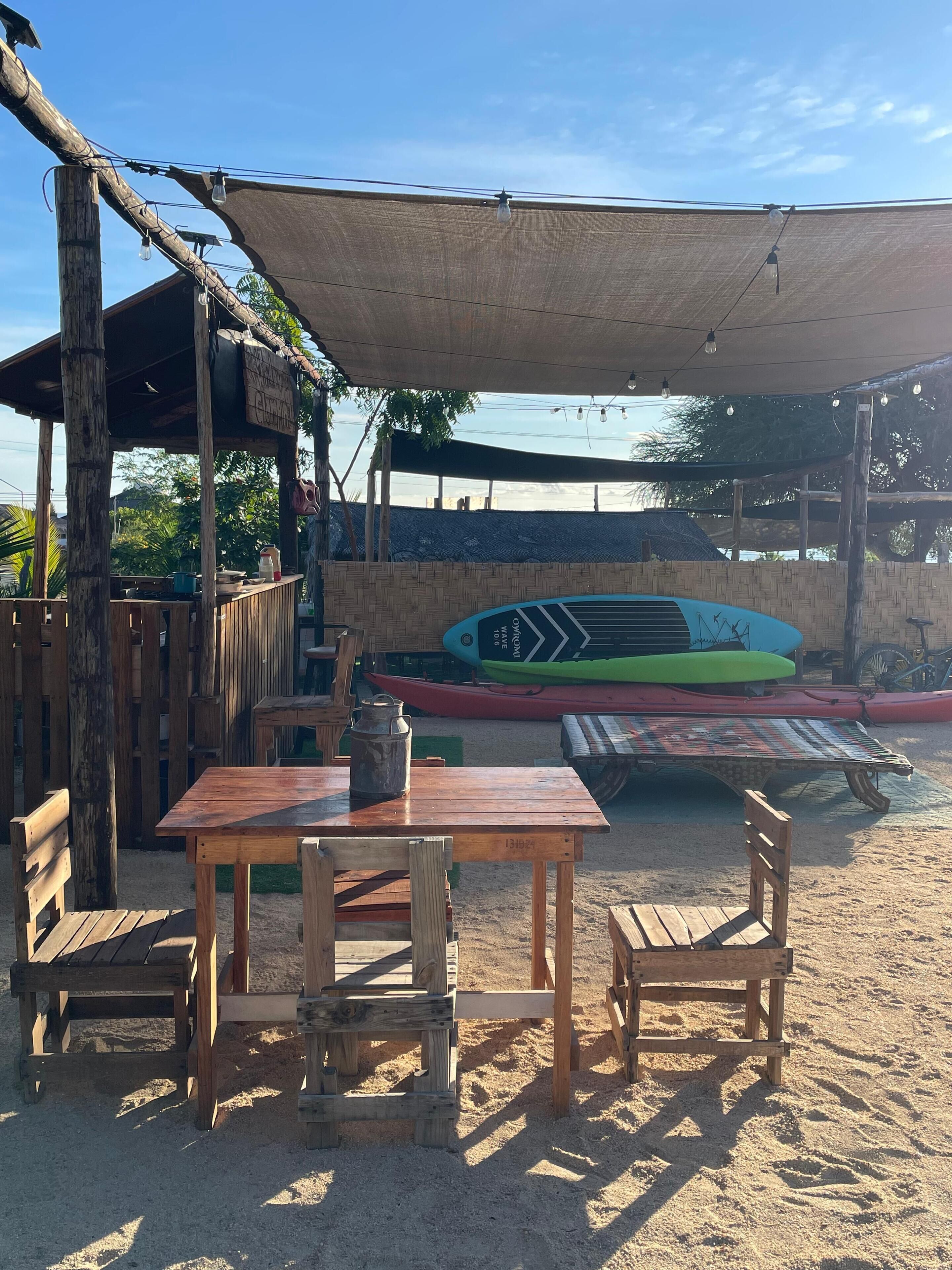 Uma área de estar externa com bancos de madeira, uma mesa, cadeiras e um toldo, com caiaques ao fundo.