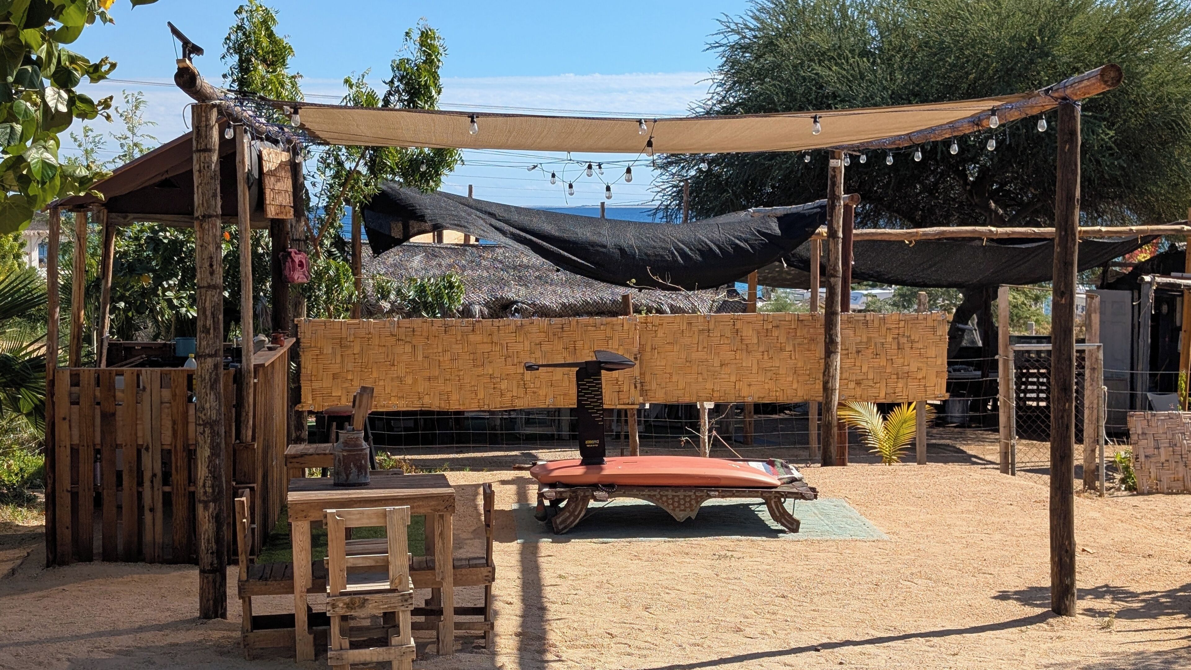 A rustic outdoor seating area with a wooden table and chairs, a thatched-roof structure, and a shaded canopy.