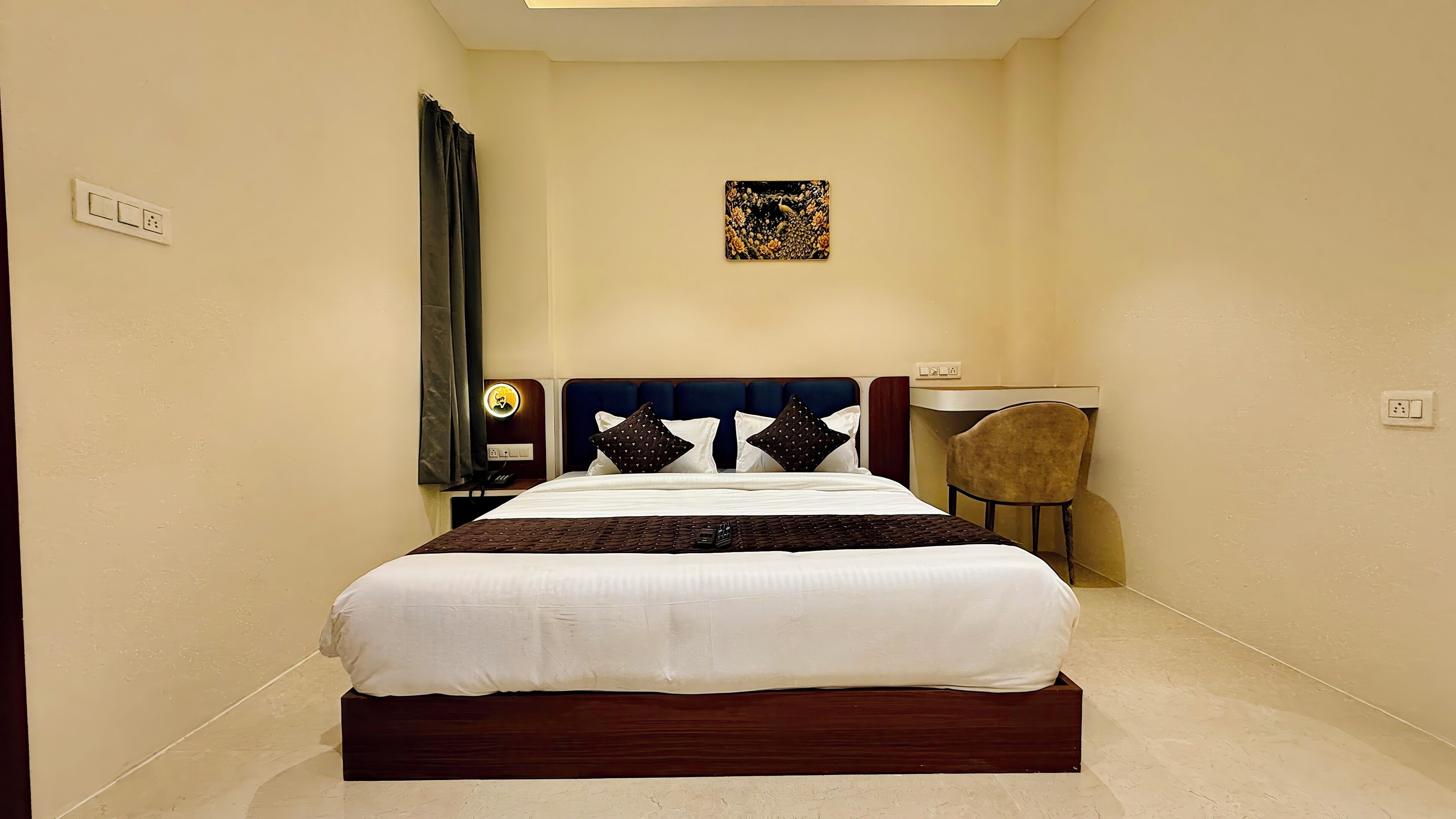 Een netjes opgemaakt bed met witte beddengoed en een donker hoofdbord, een nachtkastje met een lamp, een stoel en een klein kunstwerk aan de muur.