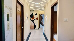 A hallway with a modern ceiling design, a wooden door with a checkmark symbol, and a wall with abstract art. - Hotel Mantri Ji (Ujjain)