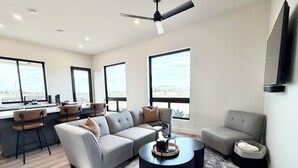 A modern living room with a grey sectional sofa, a round coffee table, and a TV mounted on the wall. There are large windows providing a view of the outdoors. - Relaxing Wine Country Stay - Hot Tub - Agave Farm (Temecula)