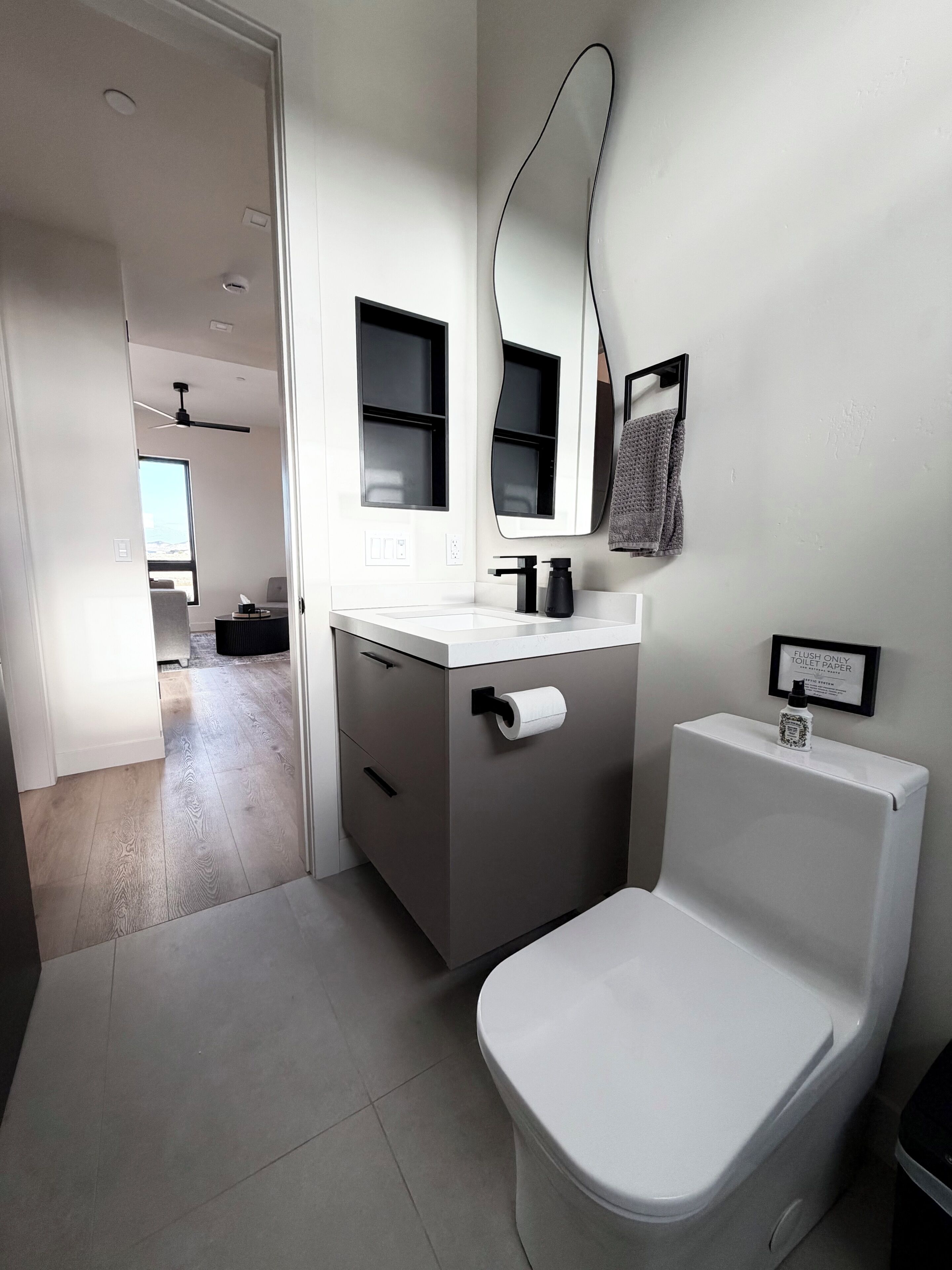 A modern bathroom with a toilet, sink, and mirror. There is a towel hanging on the wall and a sign indicating the toilet is for flushing only.