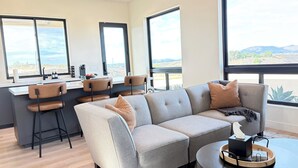 A modern living room with a grey sectional sofa, two chairs, a coffee table, and a kitchen area with a bar counter. - Relaxing Wine Country Stay - Hot Tub - Agave Farm (Temecula)