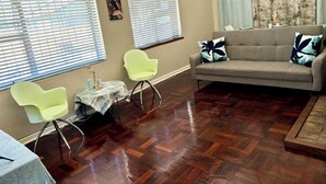 Una sala de estar con un sofá gris, dos sillones verde claro, una mesita con mantel y un piso de madera en diseño espiga.