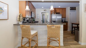A kitchen with two bar stools, wooden cabinets, a stainless steel refrigerator, and pendant lights. - Coastal Charm Awaits in Tiki Time on Topsail! (North Topsail Beach)