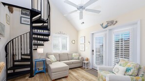 Ein Wohnzimmer mit einem beigen Sofa, einem blauen Beistelltisch, einem weißen Sessel, einer schwarzen Wendeltreppe und einem Deckenventilator.