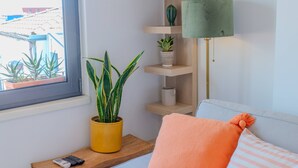 Een gezellige woonkamer met een bank, een klein tafeltje met een potplant, een lamp en een raam met uitzicht op gebouwen.