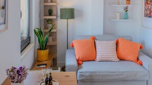 Een moderne woonkamer met een grijze bank, oranje kussens, een houten salontafel met drankjes, een gele plant en een vloerlamp.