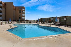 A swimming pool with lounge chairs and umbrellas, surrounded by a wooden fence and a building with balconies. - Coast is Clear– Oceanfront Escape at Topsail Dunes (North Topsail Beach)