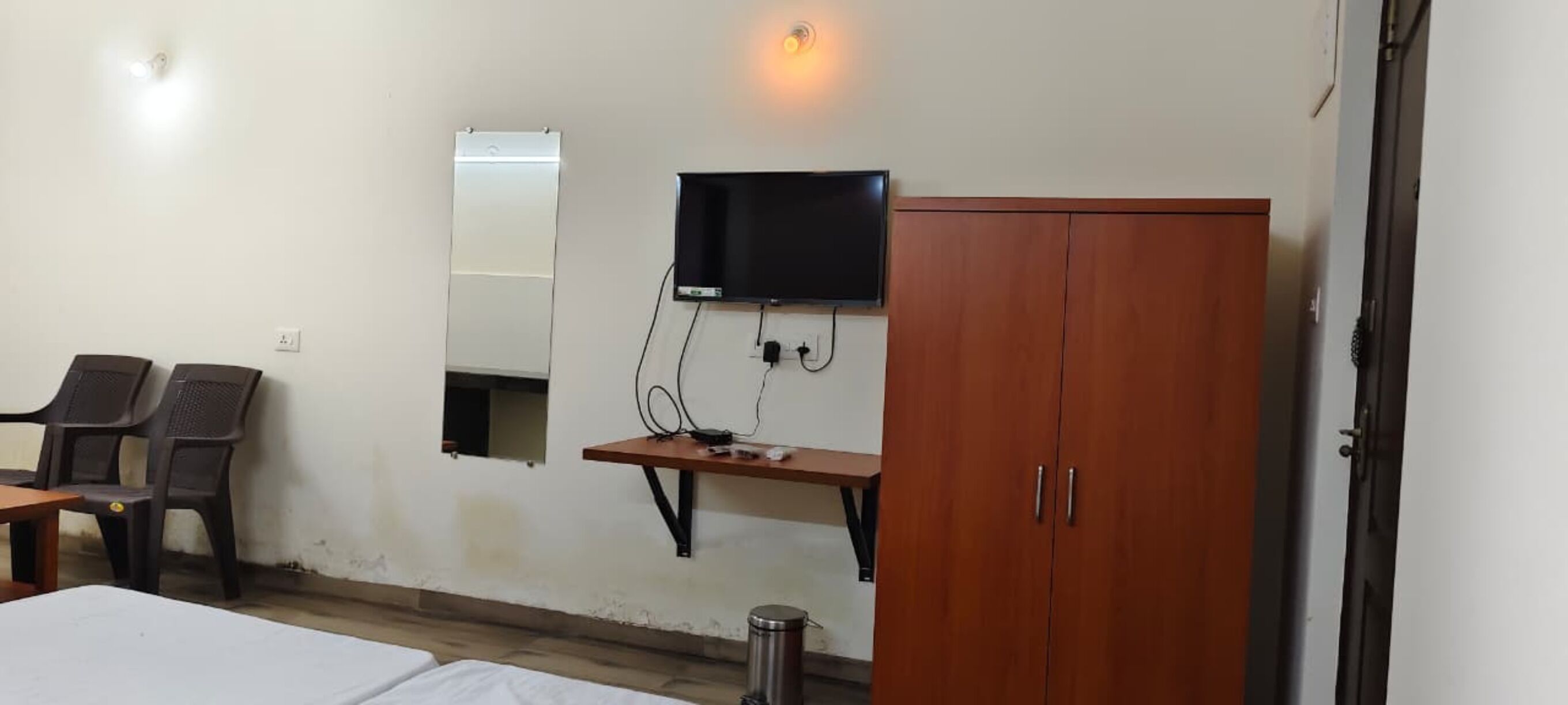 A room with a bed, a wooden wardrobe, a TV mounted on the wall, a small table, and two chairs.