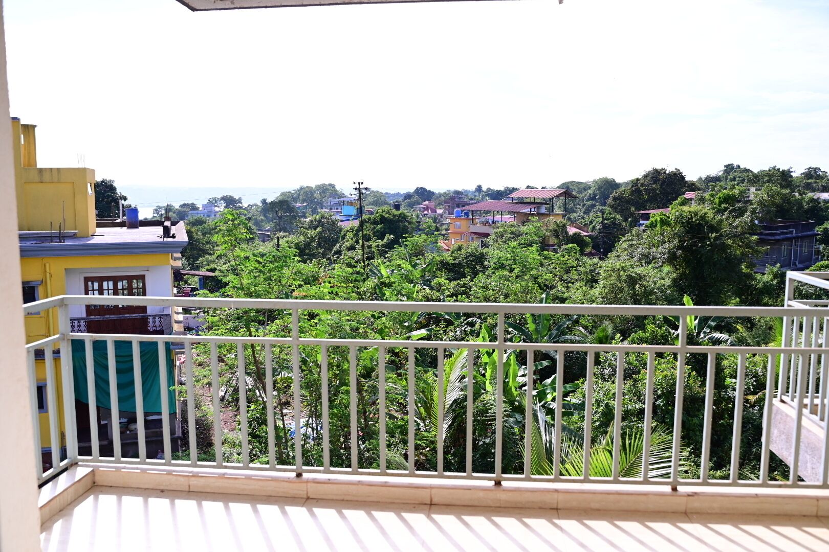 Un balcone con ringhiera bianca che si affaccia su una zona ricca di vegetazione, con edifici sullo sfondo.