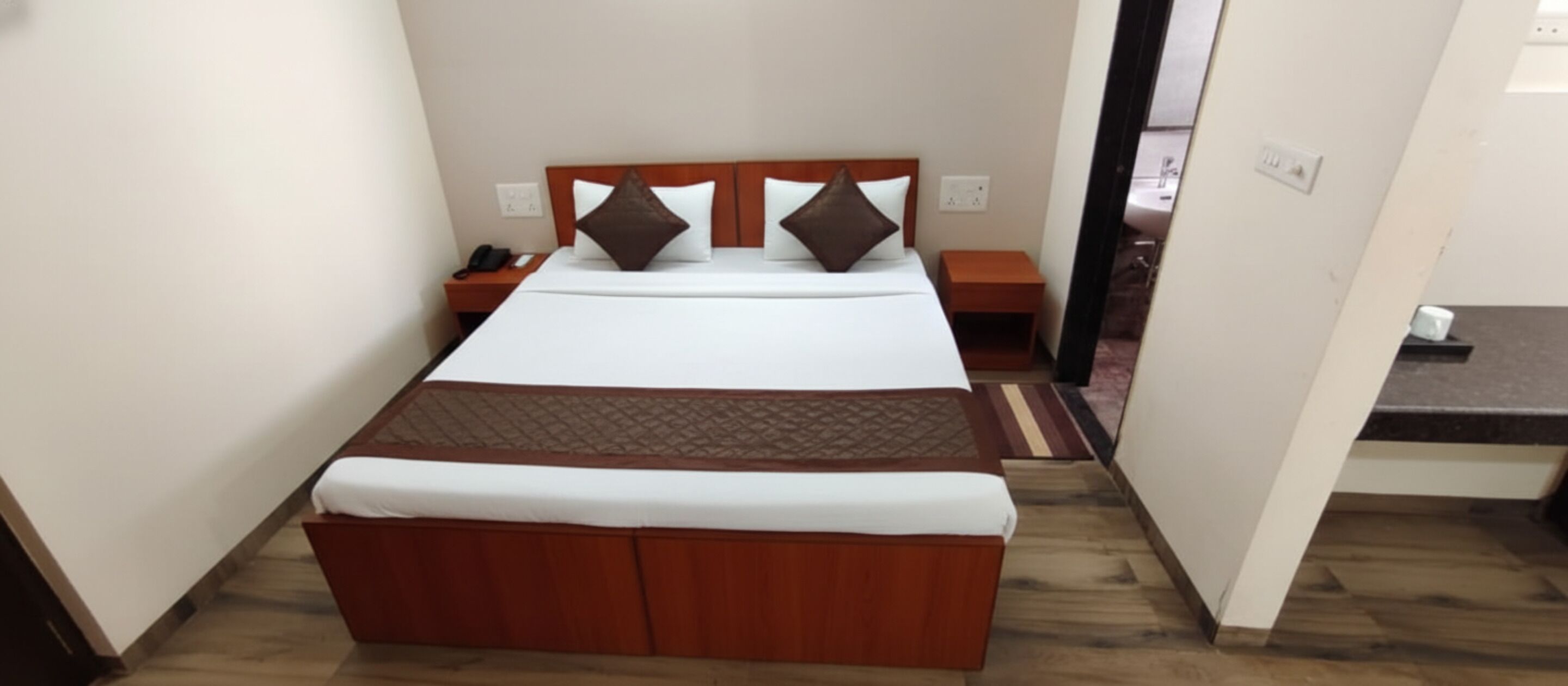 A neatly made bed with a wooden headboard, a bedside table with a phone, and a nightstand with a lamp.