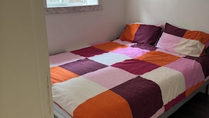 A single bed with a colorful quilt, a window with blinds, and a wooden floor. - Cedar-Cottage Comfort Suite (Vancouver)
