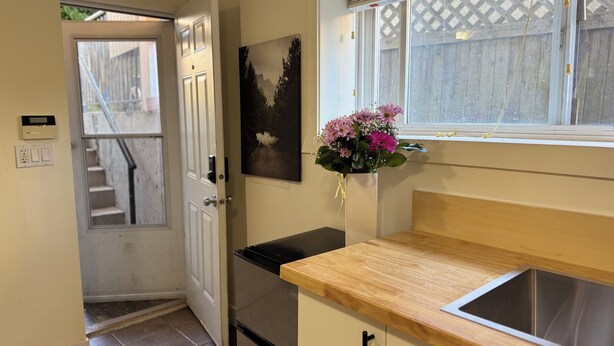 A kitchen with a wooden countertop, a stainless steel sink, a refrigerator, a door leading to a staircase, and a window with a view of a wooden fence. - Cedar-Cottage Comfort Suite (Vancouver)