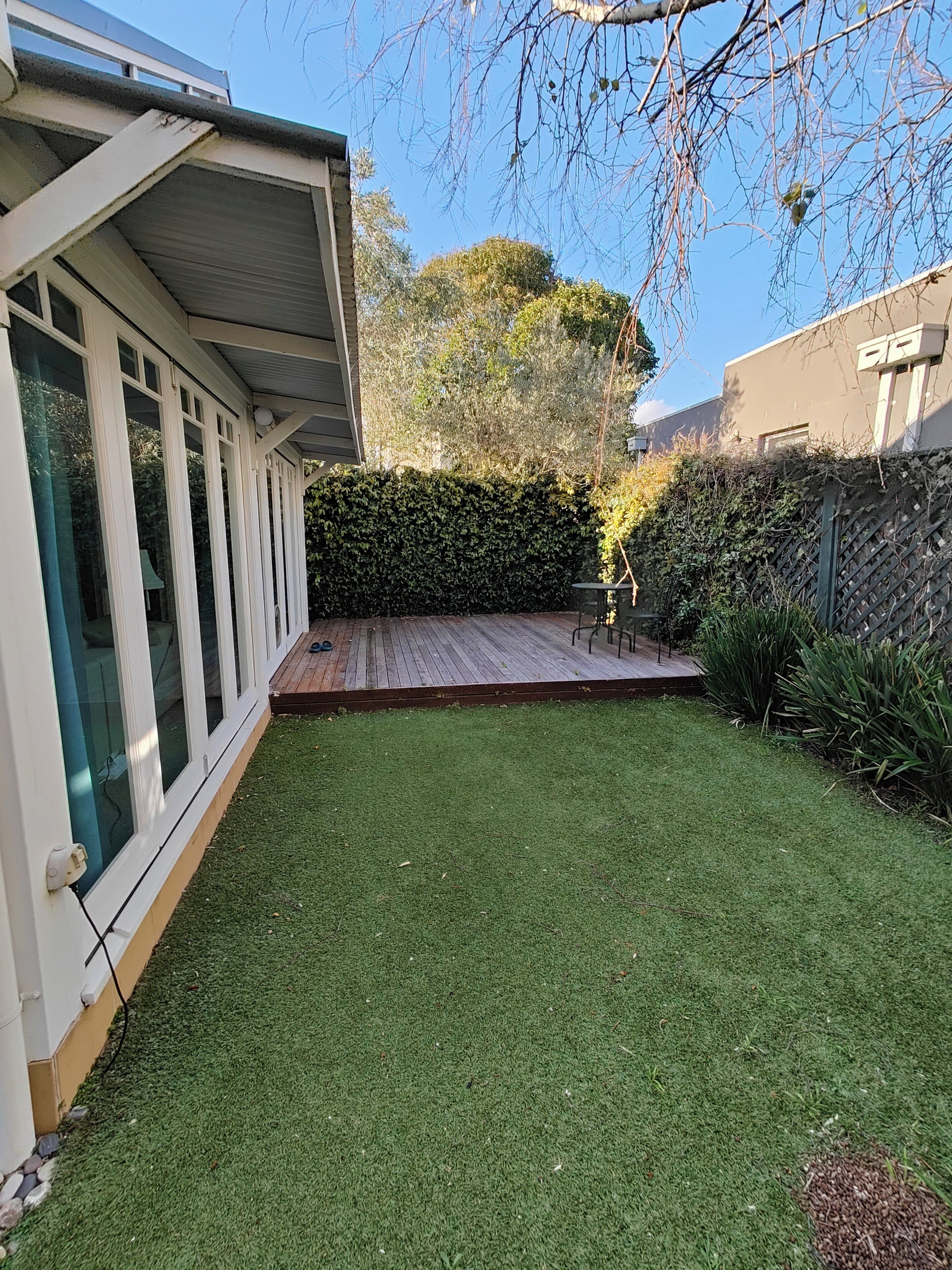 Un jardin avec une terrasse en bois, une pelouse verdoyante et une haie.