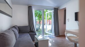 Ein modernes Wohnzimmer mit einem grauen Sofa, einem weißen Couchtisch und einer Glastür, die zu einem Außenbereich mit Bäumen führt.
