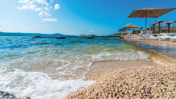 Una playa de guijarros con agua cristalina, un muelle y sillones de playa bajo sombrillas.