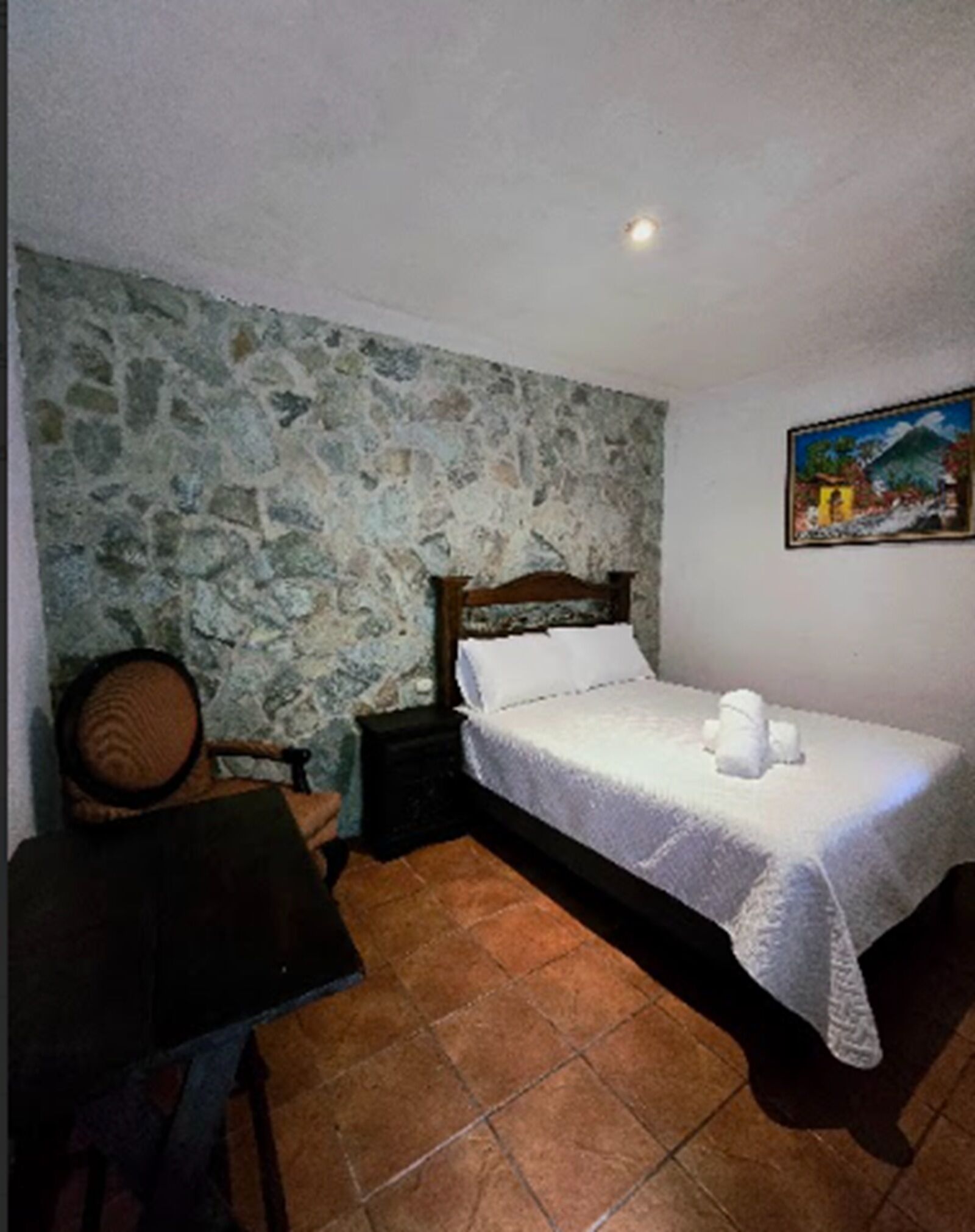 A bedroom with a stone wall, a bed with a white bedspread, a nightstand, a chair, and a painting on the wall.