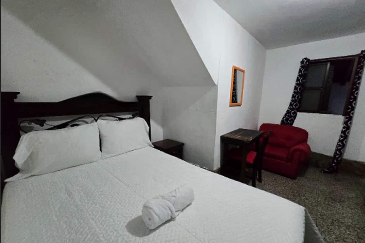 A bedroom with a bed, a nightstand, a chair, and a window with patterned curtains.
