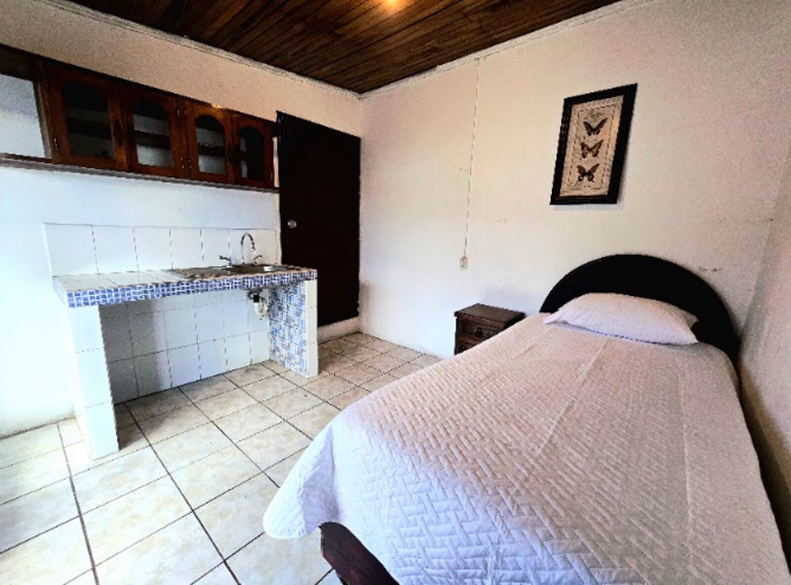 Een slaapkamer met een bed, een klein nachtkastje, en een kitchenette met een spoelbak en kastjes.