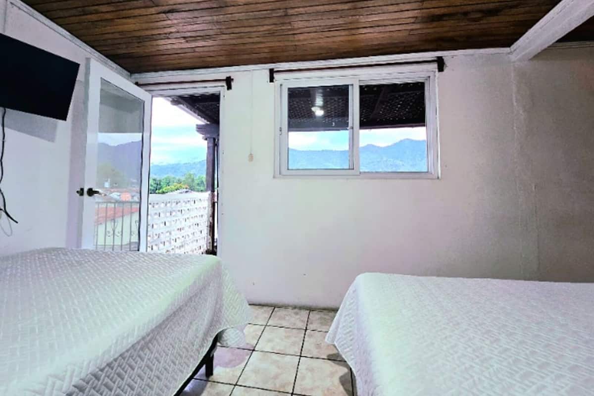 A room with two beds, a wooden ceiling, a window with a view of mountains, and a balcony with a railing.
