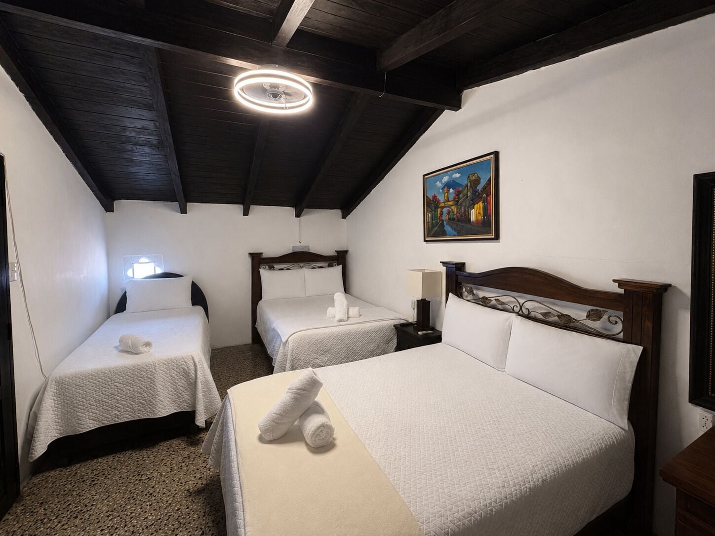 Ein Hotelzimmer mit zwei Einzelbetten, einem großen Bett, Holzbalkendecke, einem Gemälde an der Wand und einer Deckenleuchte.