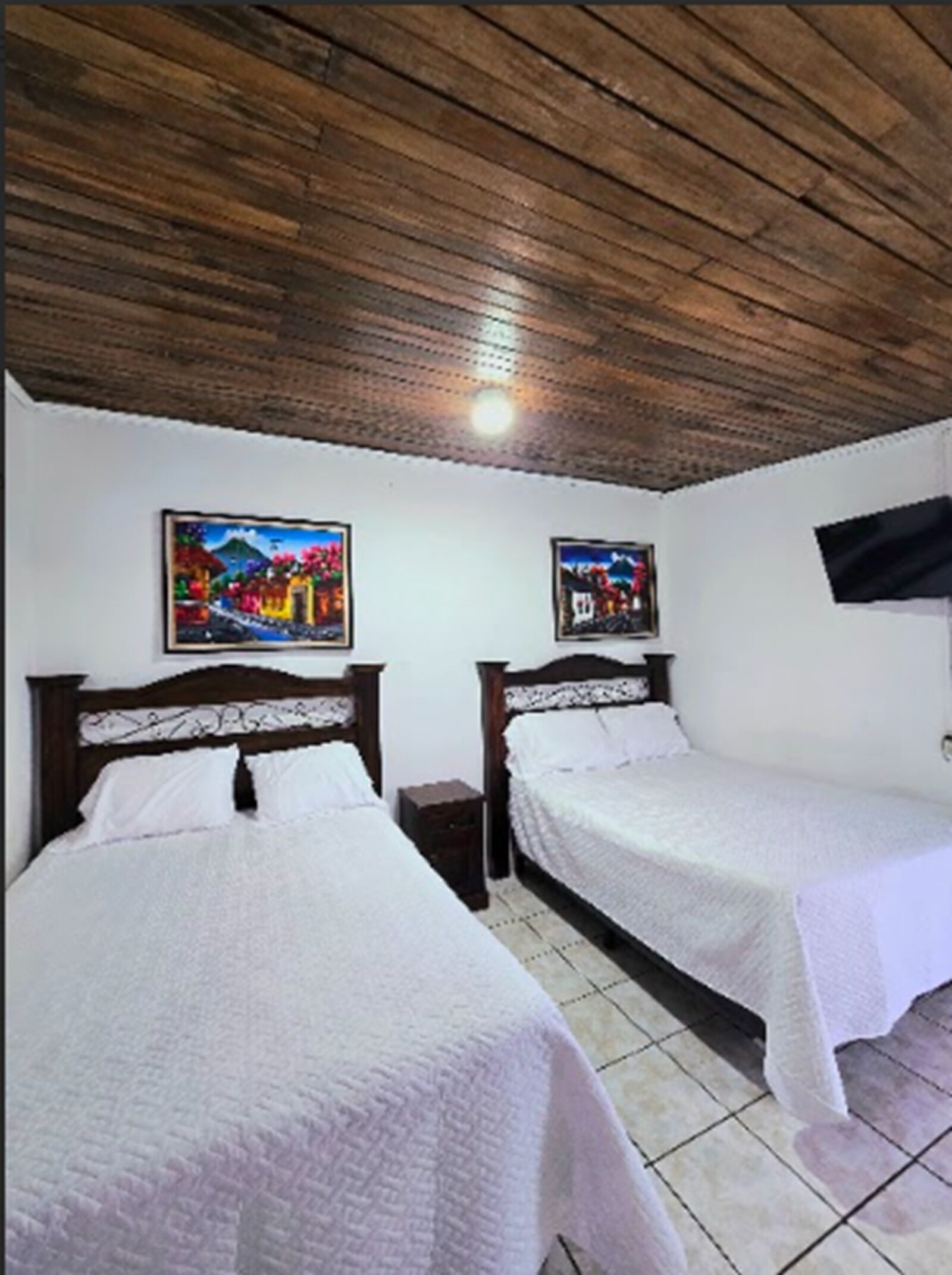 Una habitación con dos camas, cabeceros de madera, ropa de cama blanca, suelo de baldosas y un cuadro en la pared.