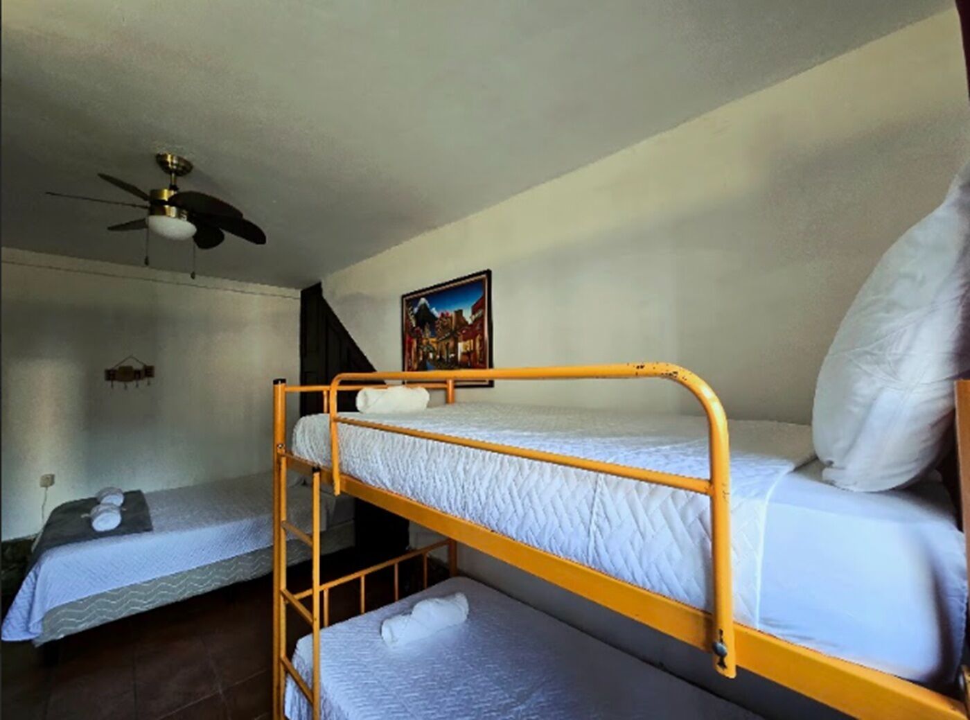 Une chambre de dortoir avec des lits superposés, un ventilateur de plafond, un tableau accroché au mur et une petite étagère.