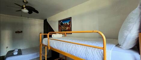 Une chambre de dortoir avec des lits superposés, un ventilateur de plafond, un tableau accroché au mur et une petite étagère.