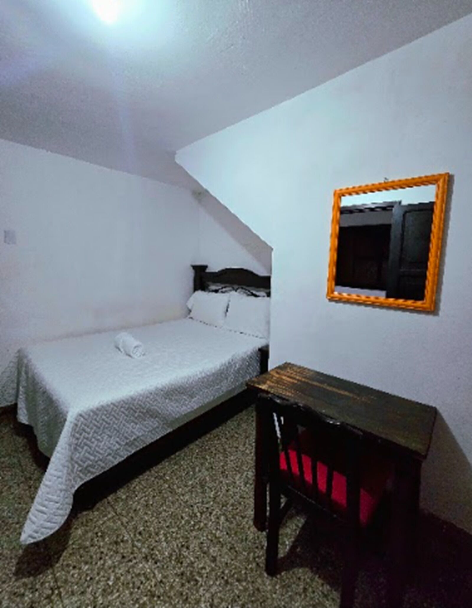 Una habitación pequeña con cama individual, una mesita sencilla y un espejo en la pared.