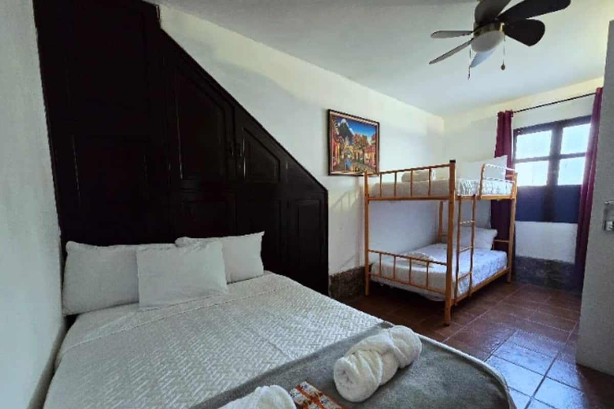 A hotel room with a bed, a bunk bed, a ceiling fan, a painting, and a window with curtains.