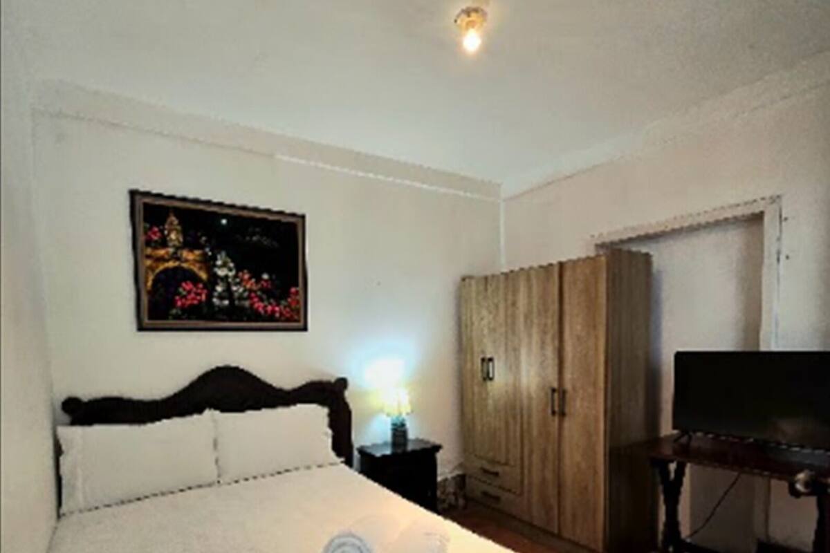 A neatly made bed with two rolled towels, a nightstand with a lamp, a wardrobe, and a television on a stand.