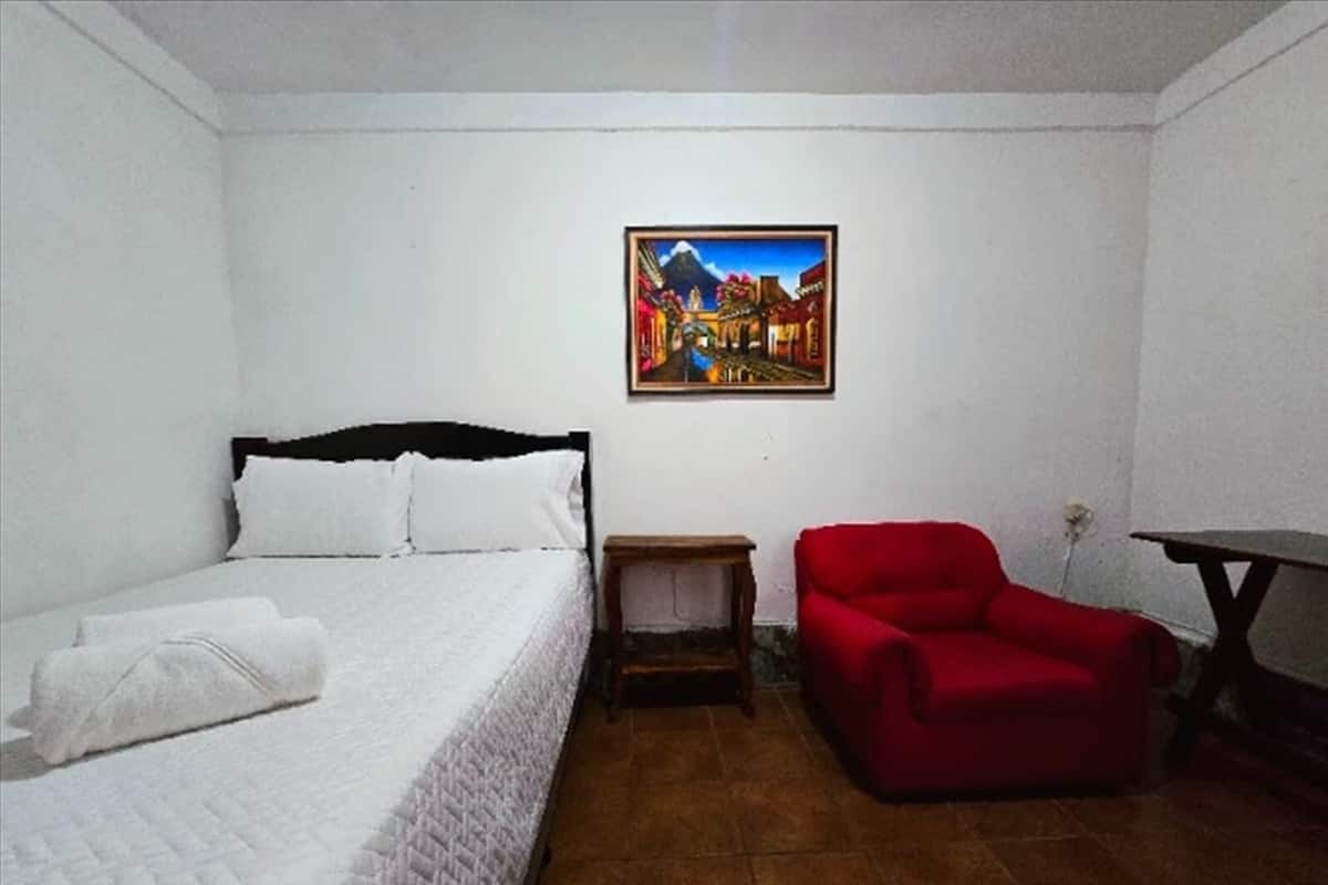 A bedroom with a bed, a red chair, a small table, and a painting on the wall.