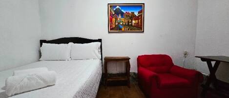 A bedroom with a bed, a red chair, a small table, and a painting on the wall.