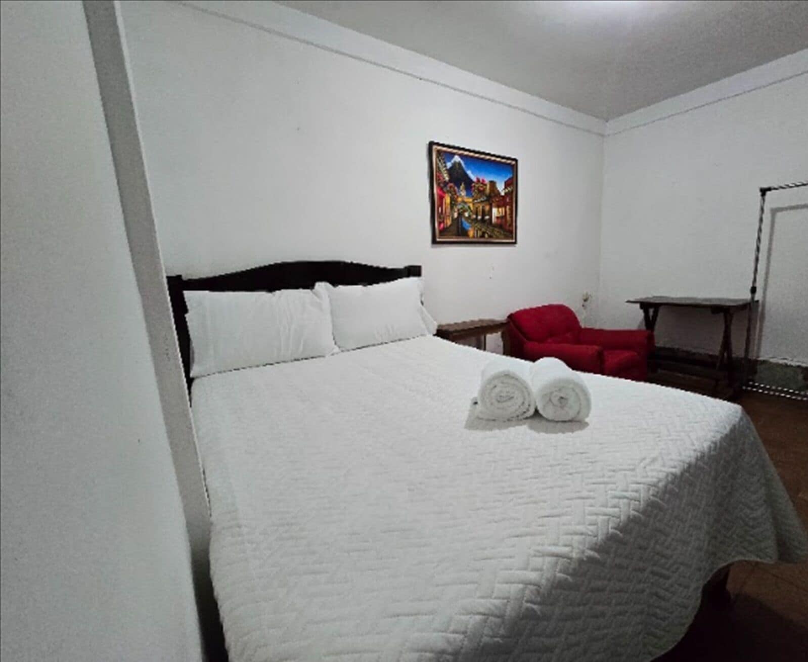 A hotel room with a bed, a painting on the wall, a red chair, and a small table.