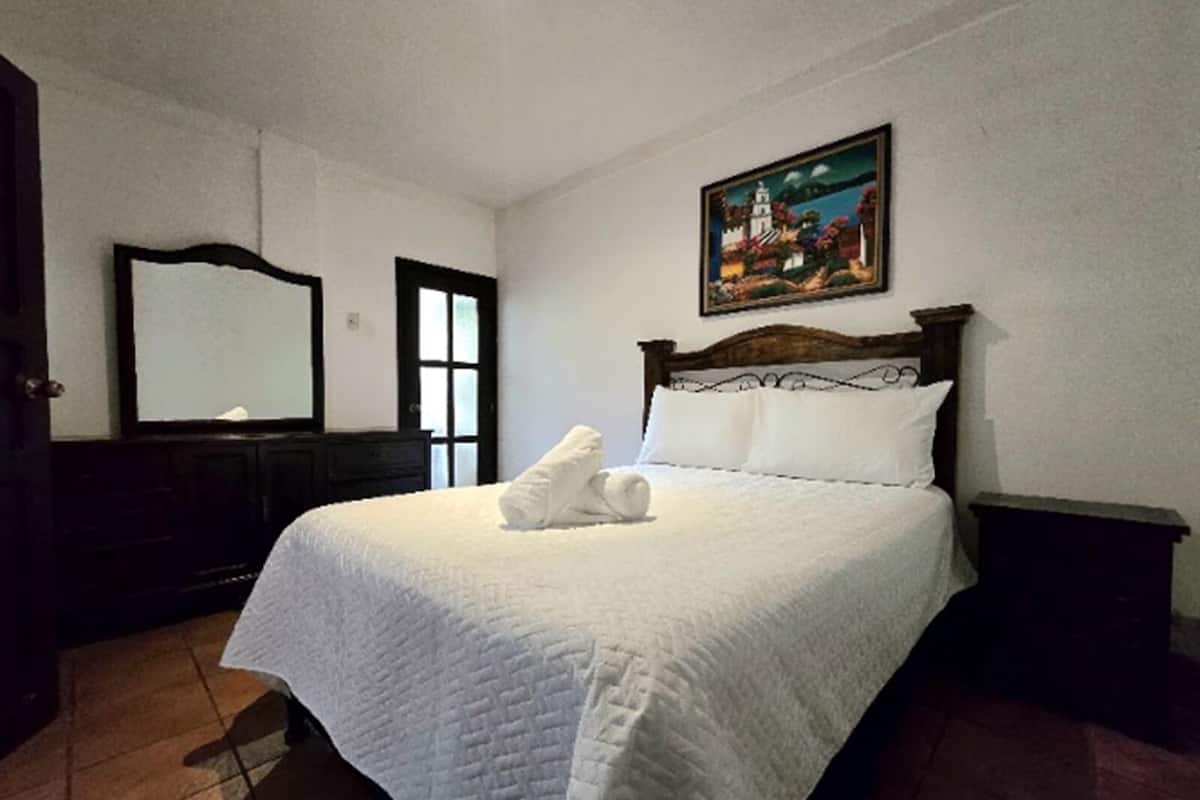 A neatly made bed with white linens and a folded towel, a wooden headboard, a painting on the wall, and a dark wooden dresser.