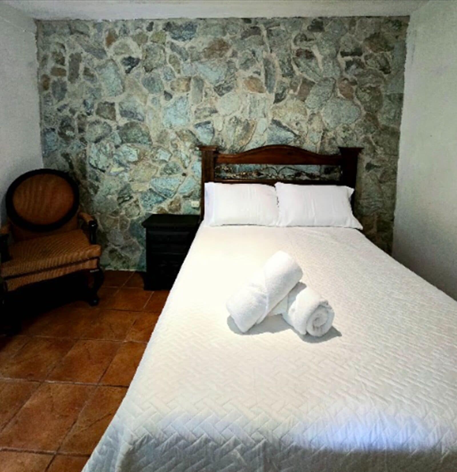 A bedroom with a stone wall, a wooden bed frame, white bedding, and a rolled towel on the bed.