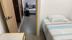Habitació petita i senzilla amb un llit, una taula i una cadira. Hi ha una porta que dona a una altra habitació.