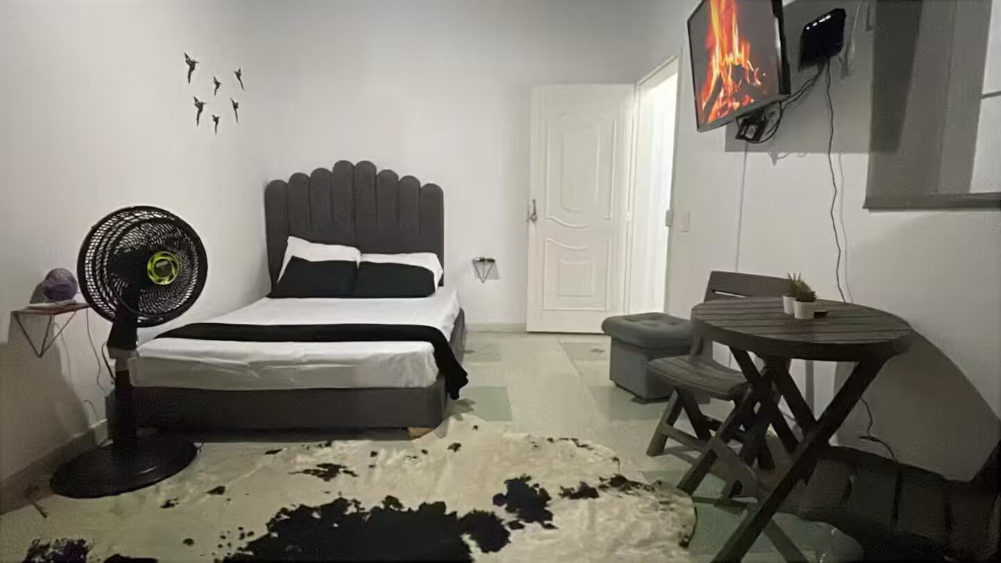 Un dormitorio con una cama, un ventilador, una mesita y un televisor montado en la pared.