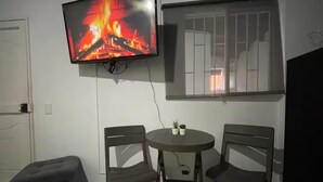 Une pièce avec un téléviseur fixé au mur qui diffuse une image de feu, deux fauteuils disposés autour d’une petite table et une fenêtre avec des barreaux.