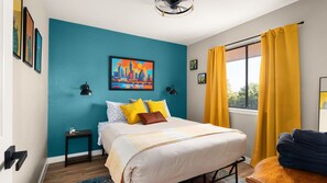 Une chambre à coucher avec un lit, une table de chevet, une fenêtre avec des rideaux jaunes, un tableau représentant un paysage urbain accroché au mur et un ventilateur de plafond.