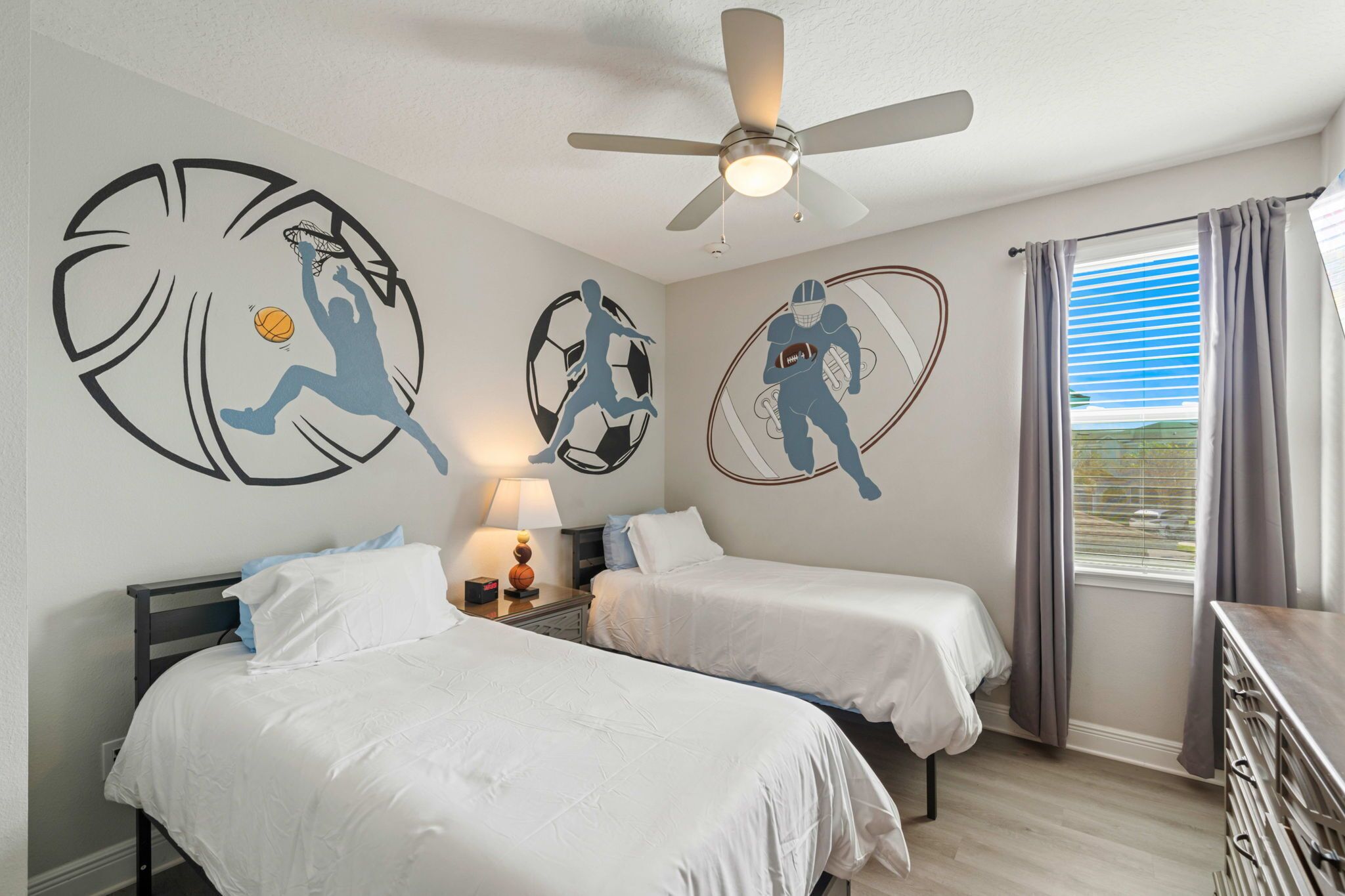 A hotel room with two beds, a ceiling fan, and wall art depicting sports themes.