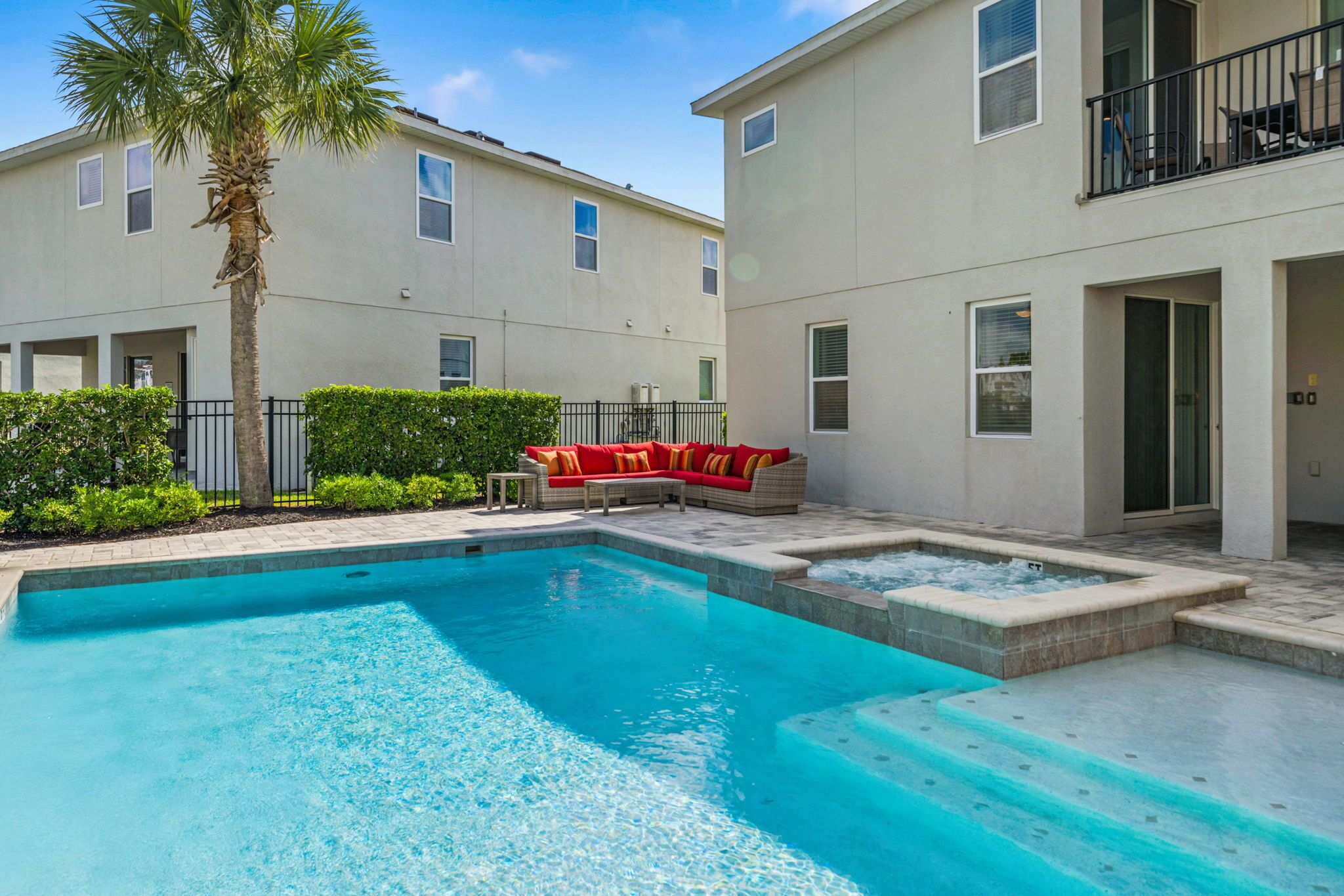 A swimming pool with a red and white outdoor seating area, a palm tree, and a two-story building with balconies.