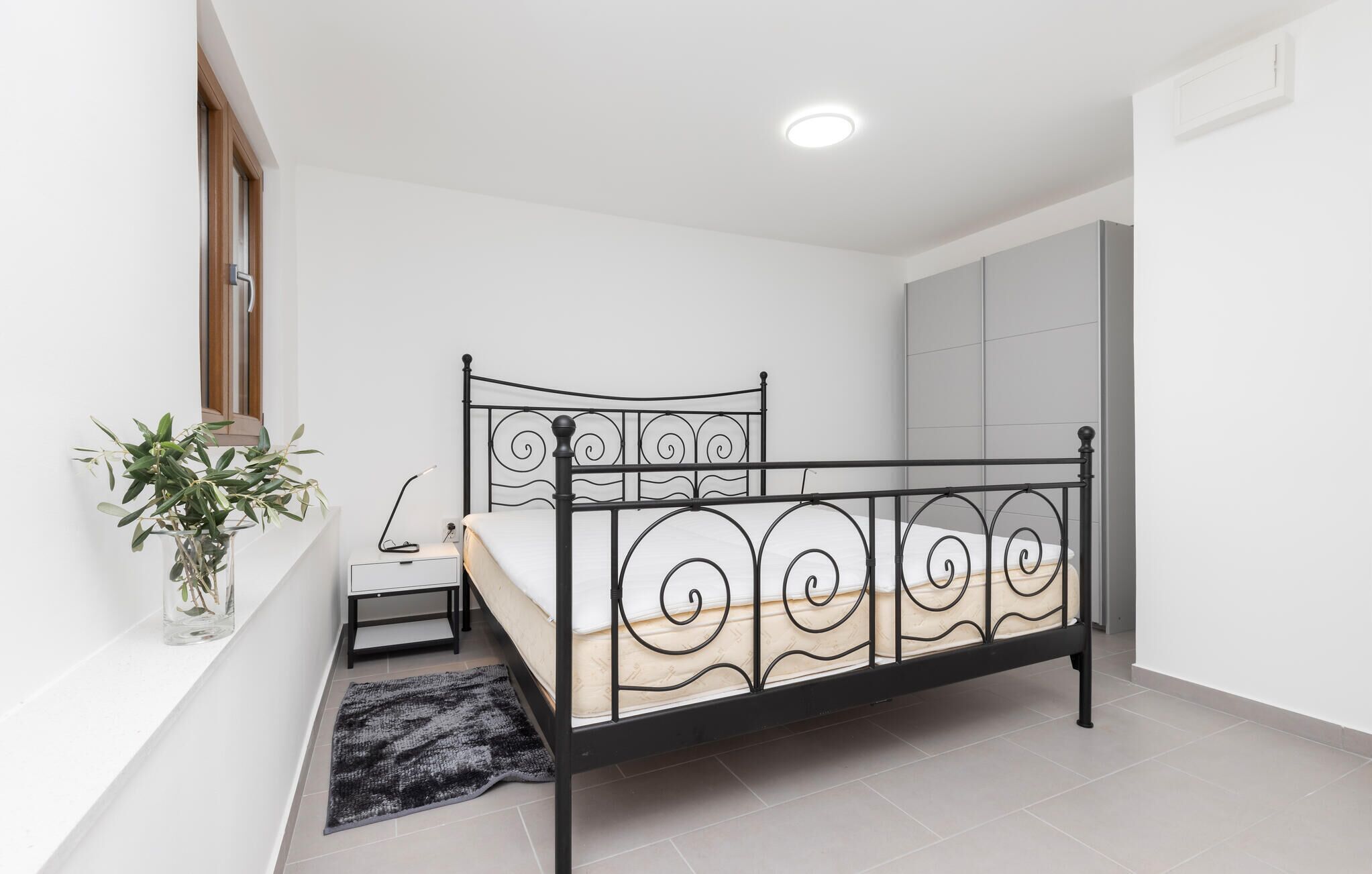 A bedroom with a black metal bed frame, a bedside table, a lamp, a potted plant, and a grey wall unit.