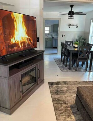 Una sala de estar con televisor mostrando una chimenea, un comedor con sillas y mesa, y una cocina al fondo.