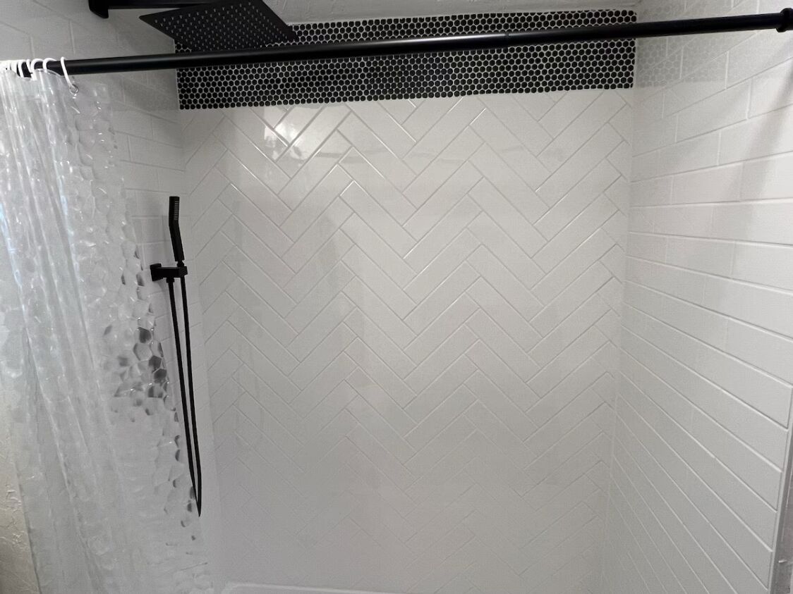 Un espace douche doté d’un pommeau de douche noir, d’un rideau de douche et d’un mur carrelé orné d’un motif en chevrons.