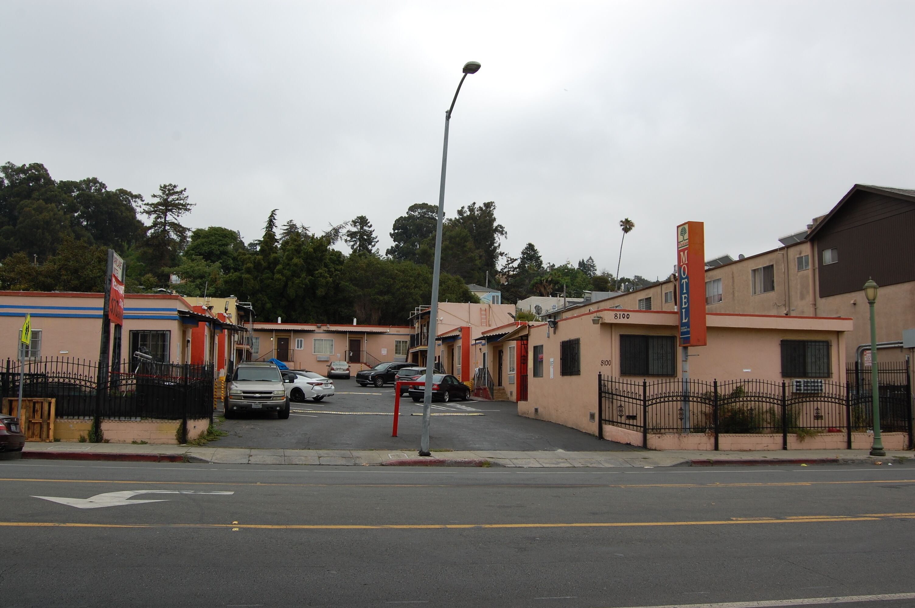 Vista de la calle con una fila de edificios, incluyendo un motel con un letrero. Hay coches estacionados a lo largo de la calle, y se ve una farola.