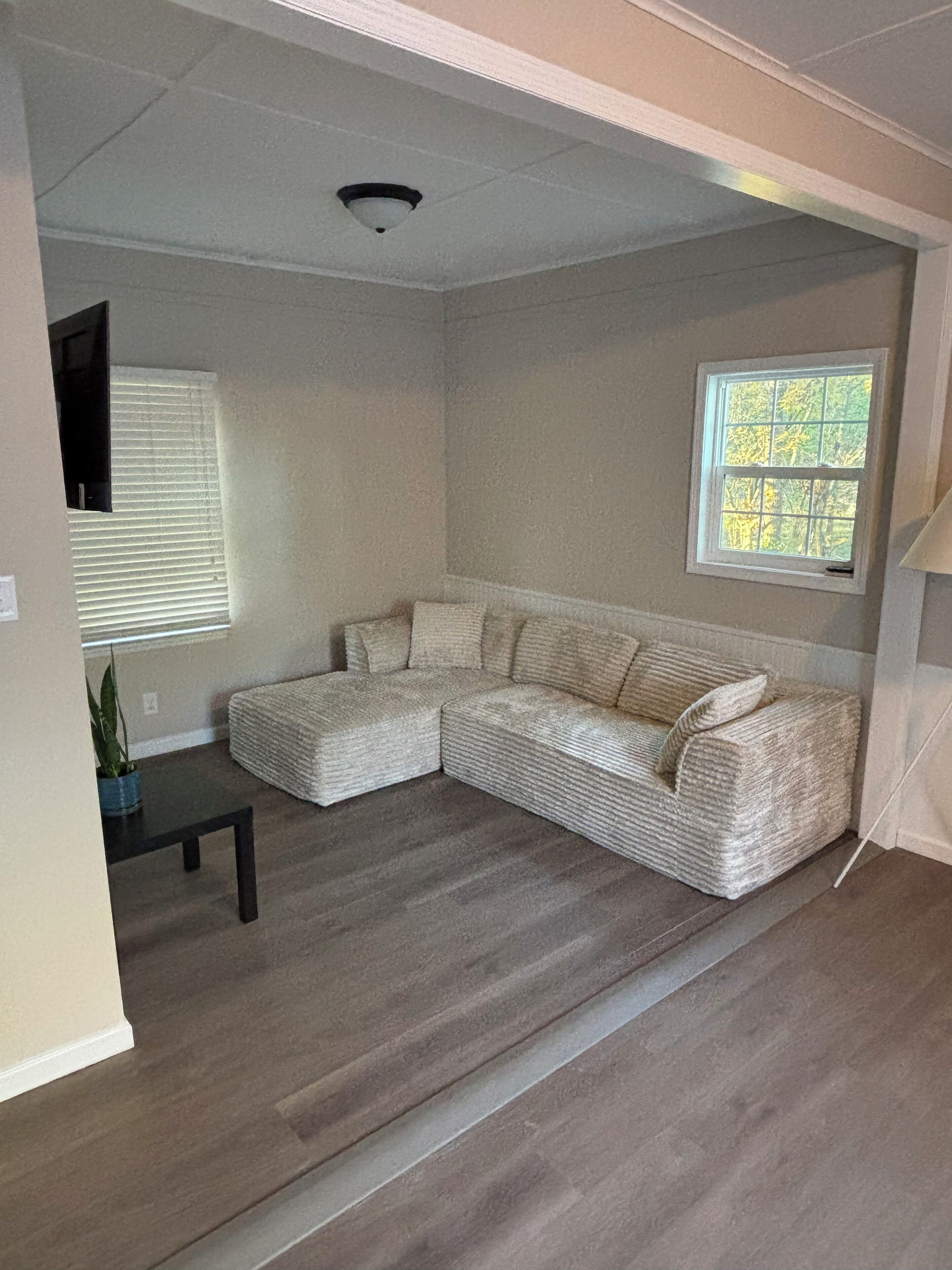 A living room with a sectional sofa, a small table, a window with blinds, and a ceiling light.