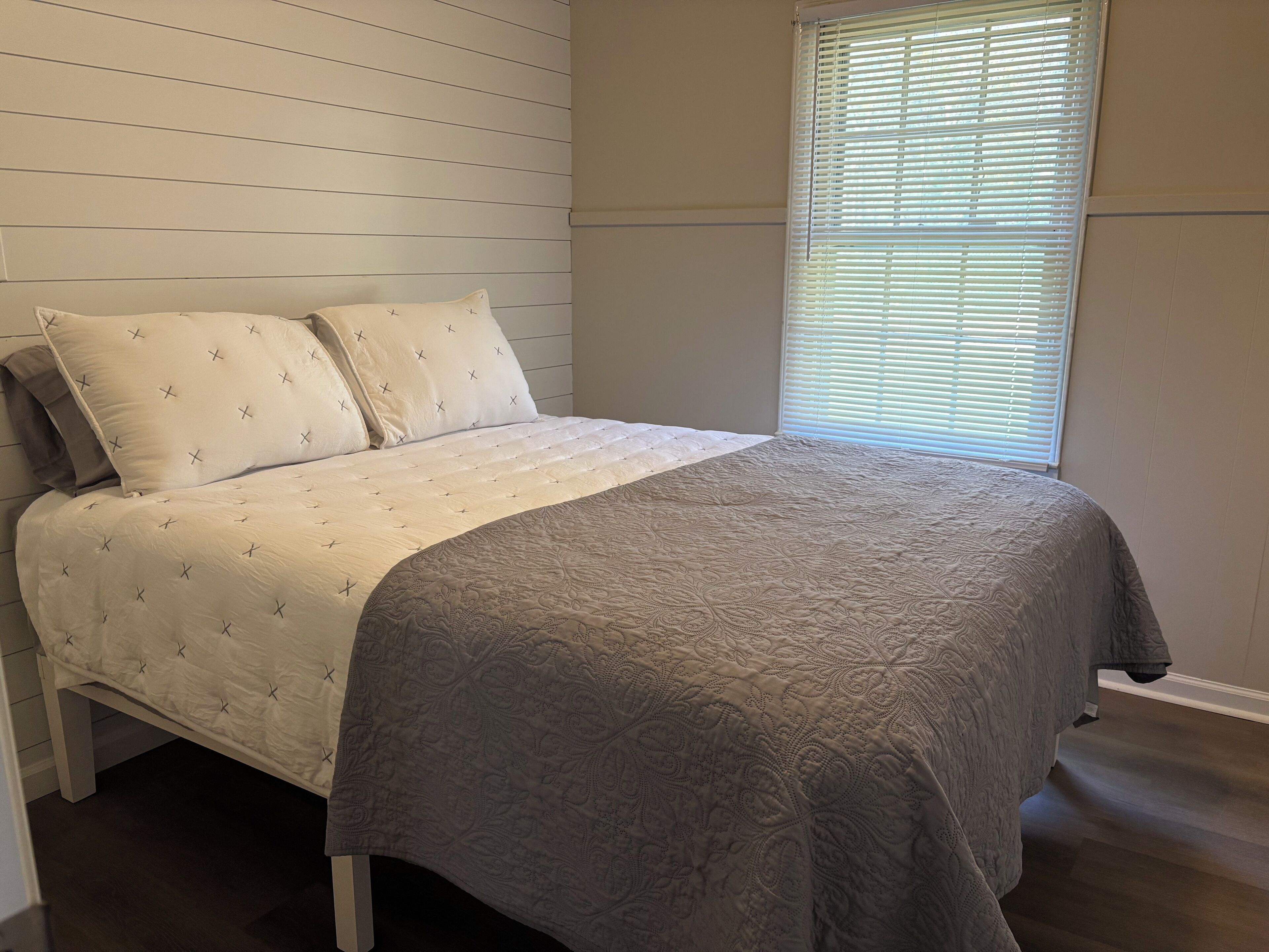 A bedroom with a bed, two pillows, a window with blinds, and a wooden wall.